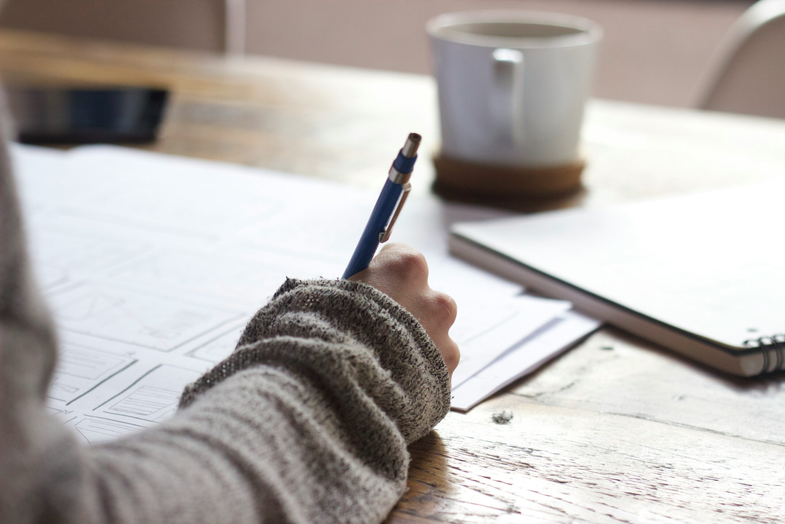 Person sitting at a desk holding a pen, writing, with a coffee cup in the background.